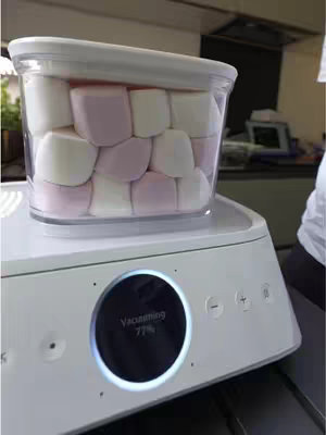 The cover image shows a close-up of a white appliance with a transparent container on top. Inside the container are a mix of white and pink marshmallows. The appliance has a black circular display showing 'Vacuuming 77%'. The background is out of focus, suggesting a kitchen setting. A white-clad figure is visible on the right side of the frame.