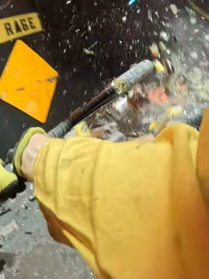 An overhead shot captures a moment of apparent destruction. A person, primarily seen by their yellow shirt and a portion of their hand in a green glove, is swinging a bat at something, causing a burst of shattered fragments. The bat is held at an angle, suggesting the impact has just occurred. In the background, a yellow sign with the word 'RAGE' is visible. The overall composition is dynamic, with motion lines from the debris and a sense of chaotic energy.