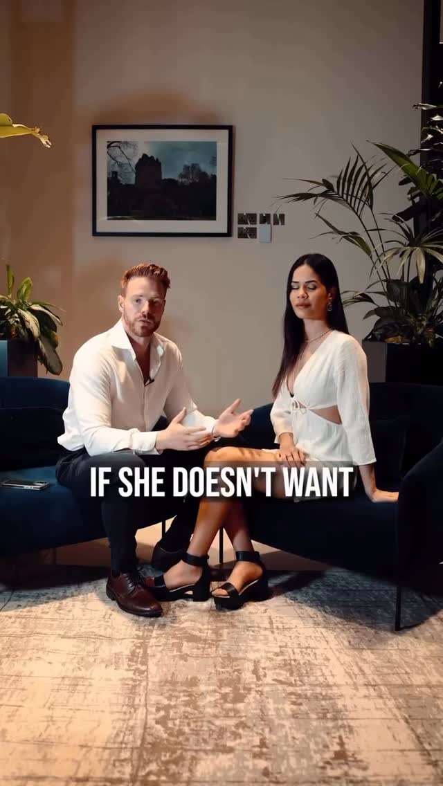 The image features a man and a woman seated on a blue couch in a well-lit setting. The man, with red hair and glasses, is gesturing expansively while speaking. The woman is seated beside him, appearing to listen or be contemplative. The text overlay reads "IF SHE DOESN'T WANT."
