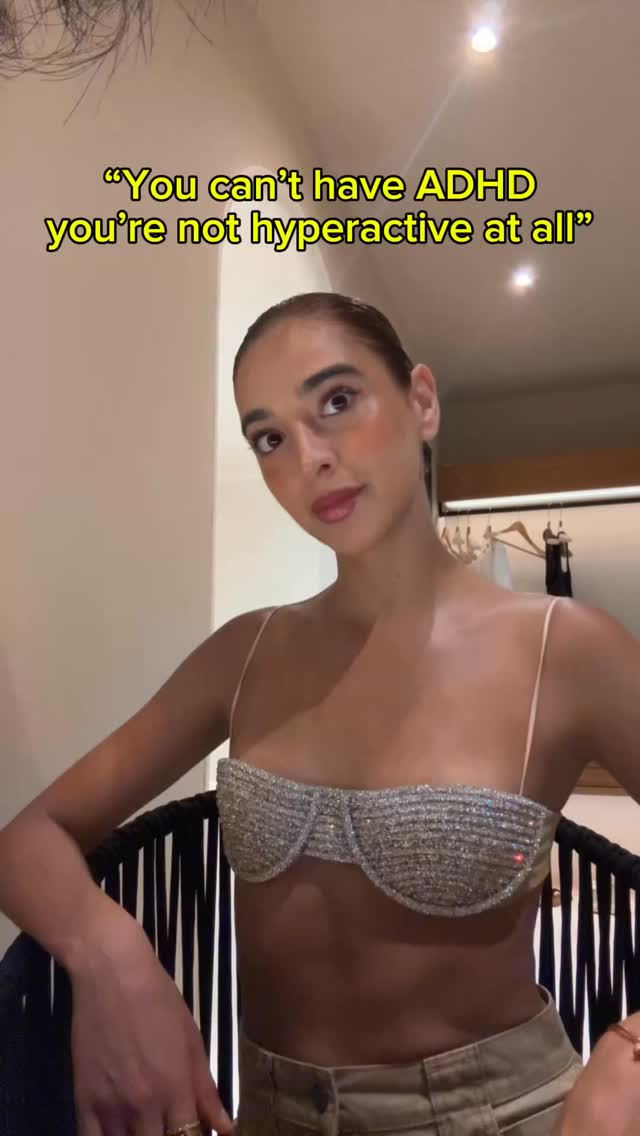 A woman with dark hair and makeup is seated, angled slightly towards the camera. She wears a sparkly top with thin straps and beige pants. The background is a neutral-toned room with some furniture visible, and an overlaid yellow text.
