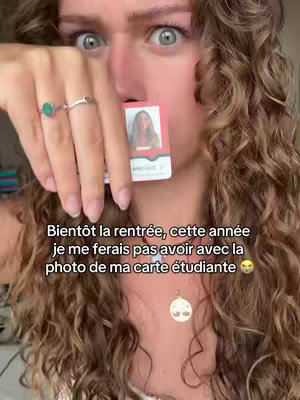 A close-up shot featuring a young woman with curly hair, wide eyes, and a surprised expression. She's holding up a student ID card to her face, partially obscuring her mouth. The background is blurred, placing focus on her and the card.