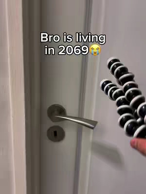 The image shows a close-up of a white door with a metal handle. Text is overlaid, humorously suggesting 'Bro is living in 2069'. A flexible tripod is visible in the foreground, held by a hand. The overall composition is simple, focusing on the door and text, with the tripod adding a modern tech element. The colors are neutral, emphasizing the contrast of the text.