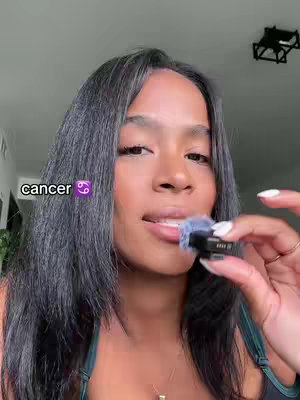 A woman with dark hair is shown in a selfie-style shot. She is holding a small device up to her mouth. The word "cancer" and a Cancer zodiac symbol appear to the left of her face.