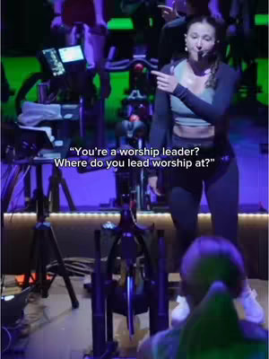 A woman in workout gear stands on a stage in front of stationary bikes. She's pointing and speaking with a microphone, likely instructing a workout class. The background has green and purple lights.