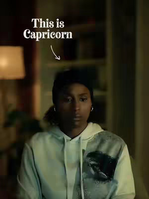 A woman with dark hair is centered in the frame, wearing a black knit cap and a light green hoodie. Above her, large, white text reads "This is Capricorn", with a white arrow pointing down towards her. The background is blurry, showing a softly lit room.
