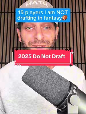 The cover image features a man with fair skin, green eyes, and short dark hair, wearing a white t-shirt and a white baseball cap with the word 'MATH' on the front. He is smiling with a microphone in the foreground. Overlayed on the image are two text boxes: a blue one that says '15 players I am NOT drafting in fantasy' with a football icon, and a red one that says '2025 Do Not Draft'. The background appears to be a vertical, wood slat wall. 
