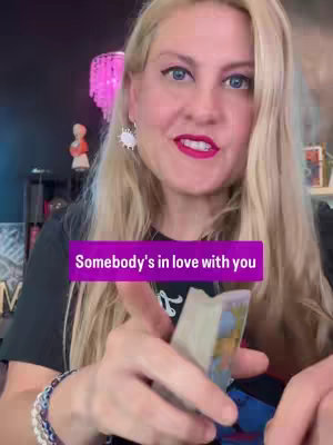 A woman with long blonde hair and blue eyes is holding a deck of cards, smiling at the camera. A text overlay reads, \"Somebody's in love with you\" in a purple rectangle.