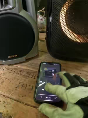 A hand in a work glove is touching a phone screen that displays a music app. The phone rests on a wooden surface, and the scene is framed with a stereo system and a speaker. The background is slightly out of focus, suggesting a casual or indoor setting. The main subjects are the phone, hand, and sound equipment.