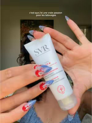 A person is holding a white tube of SVR sunscreen in front of their face. The person's well-manicured hands and nails are prominently displayed; the nails are long, pointy, and painted with a combination of red, blue, and white patterns. The background is blurred, but appears to be a room with neutral-toned decor, including a hint of a painting on the wall behind the person.