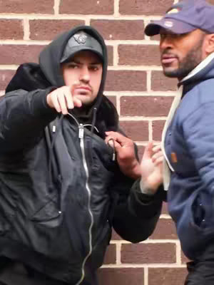 Two men are standing in front of a brick wall. One man is wearing a black zipped-up bomber jacket, a black hoodie and a cap. He is pointing his right index finger towards something and the other hand is on the other man's jacket. The second man is wearing a blue jacket and a blue cap.