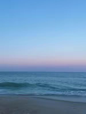 The image presents a tranquil seascape, capturing the serene beauty of the ocean and sky. The composition is divided horizontally, with the lower portion depicting the sea and sand, and the upper part displaying a gradient of blue hues transitioning into a pinkish-purple horizon. The sea's surface shows slight ripples, and the sand is smooth. The sky is clear, with no other objects in sight, giving the image a soft, calming aesthetic.