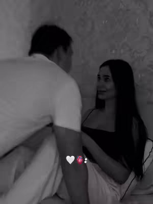 A black and white image captures a tender moment between a man and a woman, likely in a bedroom setting. The woman with long dark hair is looking up towards the man with a loving and serene expression, while he is partially turned, his face obscured. They are both in soft, casual attire. The background is intentionally blurred, putting focus on the couple, evoking a feeling of intimacy and romance. The image is further punctuated with small heart and organ emojis near the bottom, suggesting emotional bonding and affection.