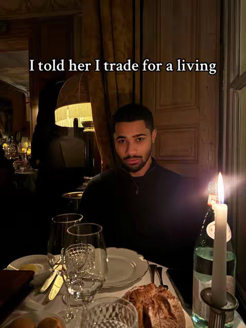 A man sits at a table in a dimly lit restaurant. There is a candle lit on the table, along with several glasses, plates, and bread. The background features curtains and a blurred view of other people and dining tables. The man is wearing a dark colored shirt and looking at the camera with a serious expression.