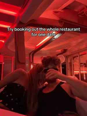 A dimly lit scene inside a vehicle or restaurant, featuring a couple seemingly on a date. The man, wearing sunglasses, is partially centered, and the woman is leaning in beside him, blurred. Red lighting and a reflective ceiling contribute to the intimate setting.
