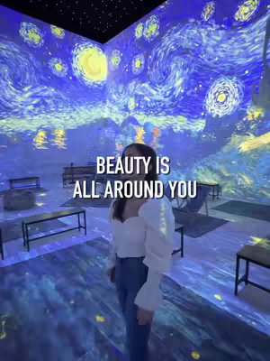 A person stands looking towards the camera.  The room is a projection of Van Gogh's Starry Night painting, with swirling blue and yellow colors covering the walls, floor, and ceiling, with tables and chairs present, but the visuals dominate.