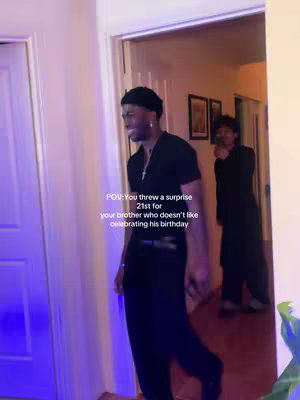 A man in a black outfit stands in a doorway, smiling and looking off-camera, with another person standing in a hallway pointing a camera behind to his back. Text overlay reads 'POV: You threw a surprise 21st for your brother who doesn't like celebrating his birthday.'