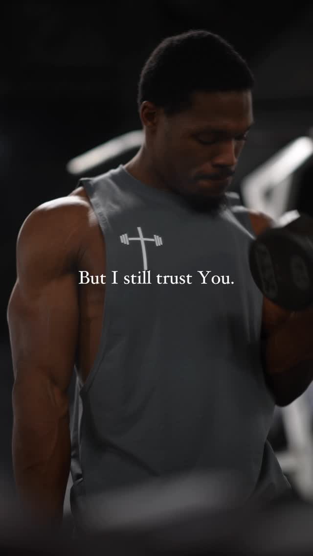 A muscular man is lifting a dumbbell in a gym setting. He is wearing a gray tank top with the image of a weight and cross. The background is dark, with gym equipment visible. Text is superimposed on the image.