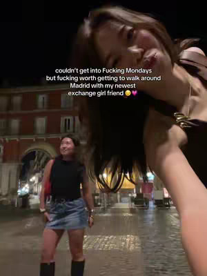 A selfie-style video taken at night in Madrid, Spain. In the foreground, a woman is filming, with her face partially visible on the right side, angled slightly towards the camera. In the background, another woman is captured on the street, slightly out of focus, with a building behind her. The scene is illuminated by streetlights, which give a slightly warm tone. The overlay text is in white and the caption expresses sentiments of embracing the moment despite the circumstances, mentioning being in Madrid with a friend.