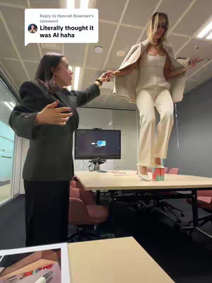 Two women in an office setting. One is standing on a table, helped by the other, who is gesturing towards her. The woman on the table holds sunglasses and is dressed in a cream-colored suit. The other woman is wearing a dark green blazer.