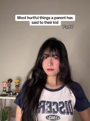A young woman with long black hair and bangs looks directly at the camera. She wears a white t-shirt with a navy collar and sleeves that has the word "DISCo" in stylized font. Behind her is a plain off-white wall, and on a shelf to her left are several small figurines of anime characters.