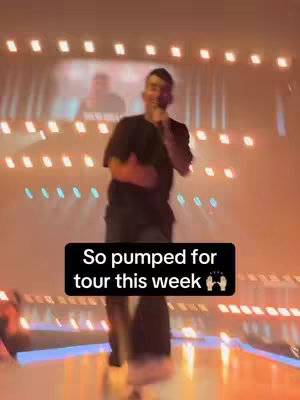 A man is on a stage with a microphone. The stage is lit with a variety of lights, and there is a screen in the background. The words "So pumped for tour this week" are displayed on a black rectangle overlay.