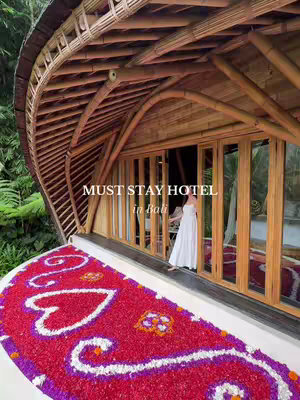 A lush outdoor scene featuring a hotel entrance with a woman standing at the doorway. The doorway is made of wooden frames and glass panels, with a red and purple flower design rug on the ground. A sign above says "Must Stay Hotel in Bali". The background is filled with thick greenery and a bamboo-style roof.