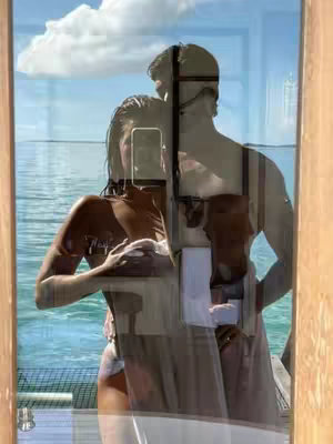 A mirror selfie captures a couple embracing outdoors. The woman, partially visible, holds the phone, while a man embraces her from behind. They stand on a wooden dock, and the reflection of the sea, sky, and clouds are visible through the glass. The overall colors are natural, with a blue sky and sea complementing the skin tones reflected.