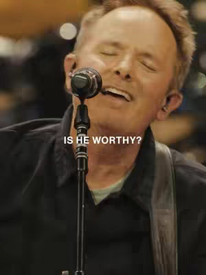 A medium shot of a man singing into a microphone, with a black microphone stand in the foreground and blurry background. The text "IS HE WORTHY?" is overlaid on the image. The man is wearing a black shirt and has his eyes closed and mouth open as if singing.