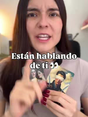 A woman with long brown hair is centered in the frame, facing the camera with a slight smile. She's holding two tarot cards, one featuring three women, and another with a man blindfolded and holding swords. Text overlayed on top reads "Están hablando de ti 👀‼️" The background is softly blurred, showing a glimpse of a room with a shelf.