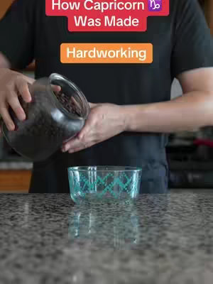 A person is seen pouring small, dark-colored objects (possibly beans) from a clear jar into a clear glass bowl with a teal geometric pattern. The person is wearing a black shirt and is positioned in front of a speckled countertop. Above the person, there's a red banner with the text 'How Capricorn ♑ Was Made', and below it, an orange banner reading 'Hardworking.'