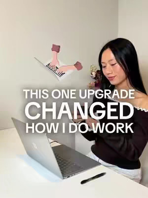 A woman with long black hair is seated at a white desk, angled towards a laptop that sits open in front of her. The background is a clean, white wall with a stylized illustration of hands appearing to type on a laptop keyboard. Overlaid on the image is large text that reads 'THIS ONE UPGRADE CHANGED HOW I DO WORK.'