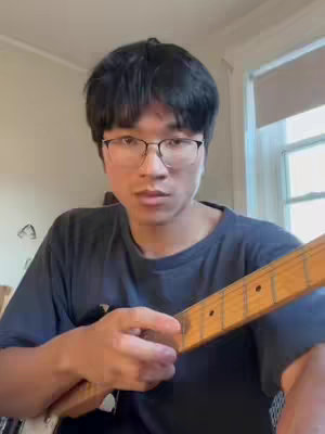 A medium shot of a young man with black hair and glasses, looking towards the camera while holding a guitar. The background is a room with a window and wall.