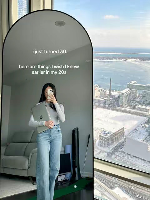 A woman stands in front of a large, arched mirror, holding a laptop and a phone. The mirror's reflection shows a city view outside a window. Text overlays the mirror, with sentences about turning 30 and the things the creator wishes she knew in her 20s. The background is a bright, modern living space next to a window.