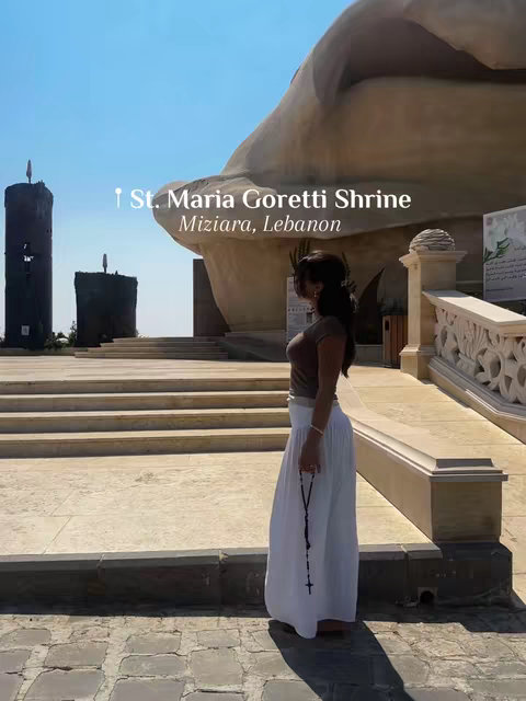 A woman in a brown top and white skirt stands with a rosary in front of a large, abstract religious structure. The sky is clear blue and the scene is bathed in natural light.