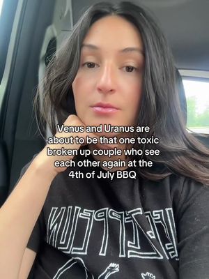 A selfie-style shot featuring a woman in a car. The woman has long dark hair and is wearing a black t-shirt with a white and black design. Overlayed text in the image offers astrological commentary. She's looking at the camera with a slight expression. The background suggests an outdoor setting, possibly a car. The lighting appears to be from the interior and exterior.