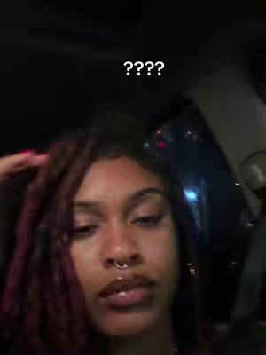 A selfie-style shot taken from inside a vehicle captures a woman with dark curly shoulder-length hair, a nose ring and glossy lips. The background is dark, with only some illumination visible near the upper right suggesting an interior or passing cityscape. The image is sharply focused on the woman's face, highlighting her expression.