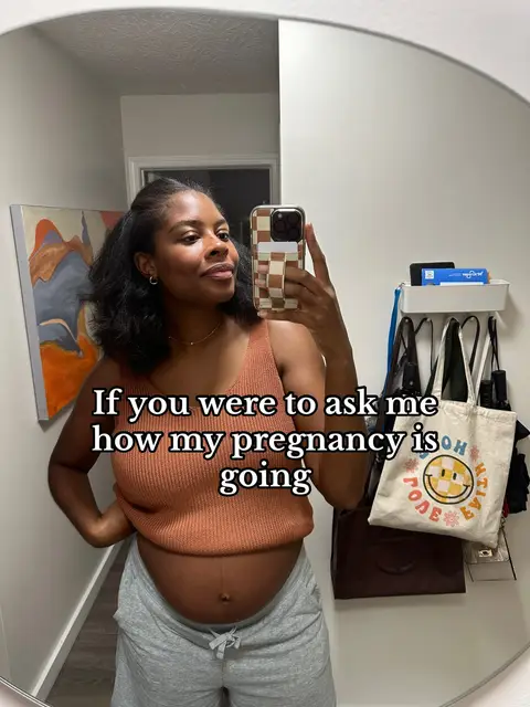 A mirror selfie shows a pregnant woman in a bathroom. She is wearing a tan top and gray sweatpants. The text overlay says 'If you were to ask me how my pregnancy is going.'