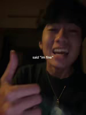 A selfie-style shot where a person is seen from the chest up. The person, who appears to be male, has dark hair and is positioned slightly off-center to the right. The background is dimly lit, suggesting an indoor setting. They are smiling and giving a thumbs-up. The text overlay in white font, "said 'im fine'," is centrally positioned near the bottom of the frame. The lighting is average, and creates a warm tone.