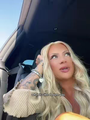 A woman with long blonde hair and heavy makeup sits in the driver's seat of a car. The interior of the car is dark.