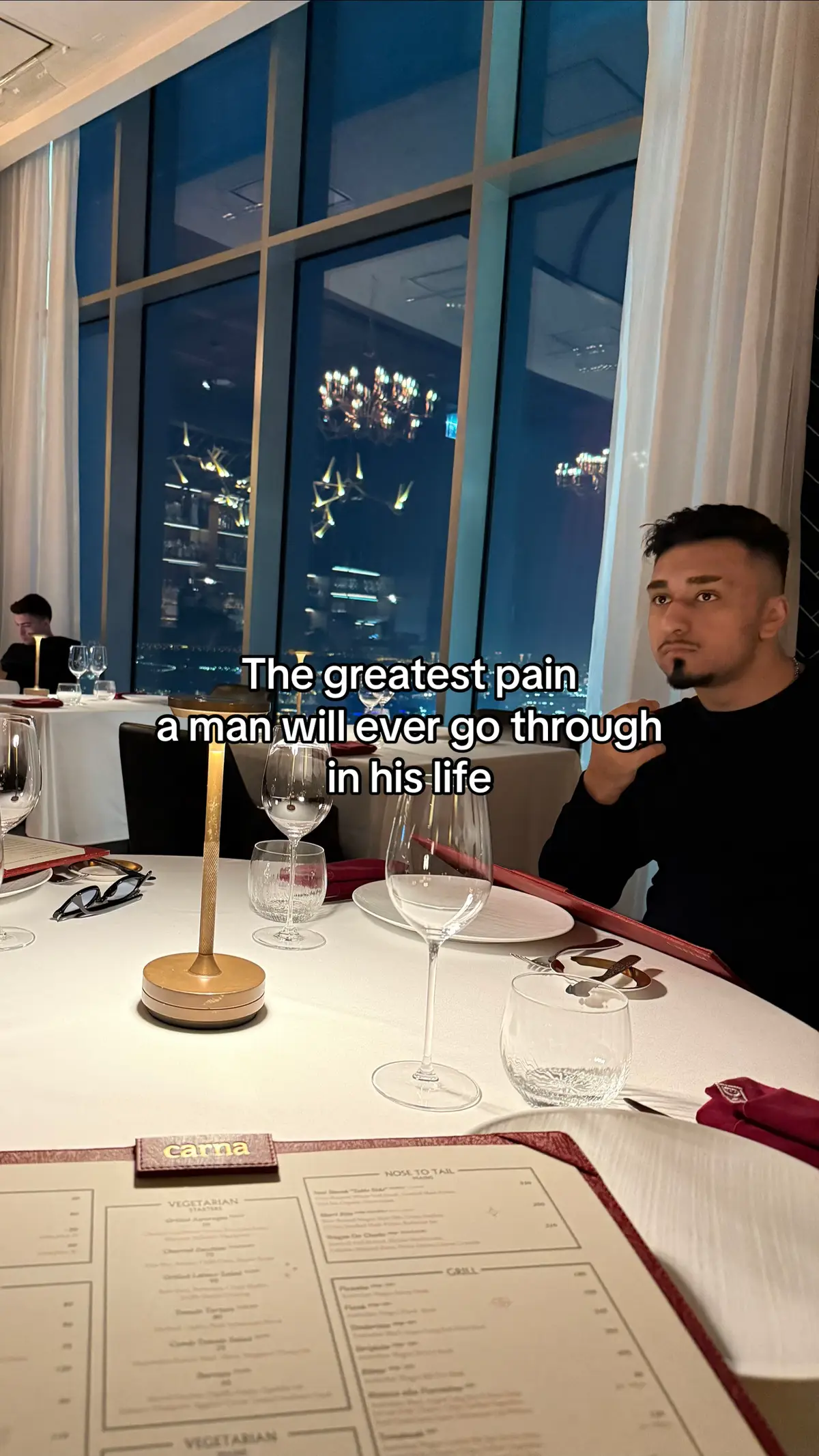 A man is seated at a restaurant table with a white tablecloth. A menu is open in front of him and a hook text overlay indicating 'The greatest pain a man will ever go through in his life'. There are wine glasses, a candle holder, and a dark view through a large window in the background.