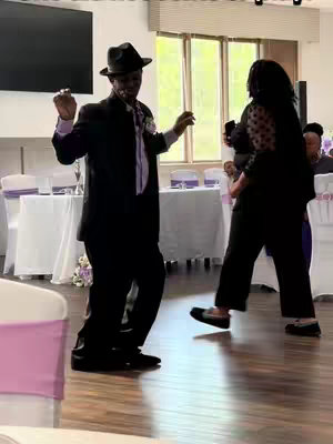 A man in a tuxedo is dancing at a wedding reception. A woman in a black top dances to the right, while other tables and chairs are in the background.