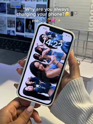 A person's hands hold a smartphone, that displays a lock screen wallpaper featuring several images of what appears to be a group of female figures, with the phone charging. The background subtly suggests an interior setting, with details like a laptop screen visible behind the phone holder and the charging cable plugged in at the bottom of the phone. Adding to the theme are some of the background items.