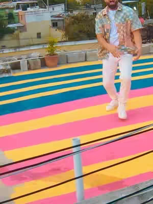 The image shows a man on a colorful rooftop. The floor is striped with pink, yellow, and turquoise. Buildings and a small potted plant are in the background. The man is wearing a patterned shirt, white pants, and white shoes. He is singing and has his mouth open.