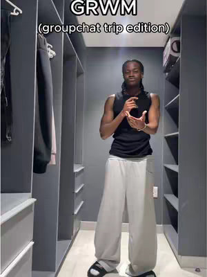 A man stands in a walk-in closet facing the camera, hands clasped near his chest. He's wearing a black tank top, light gray wide-leg pants, and black sandals with white soles. The closet has gray shelving, and the lighting is somewhat dim.