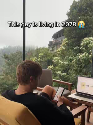 A man sits comfortably in a chair with a laptop, phone, and a scenic view in the background. Overlaid text reads, "This guy is living in 2078".