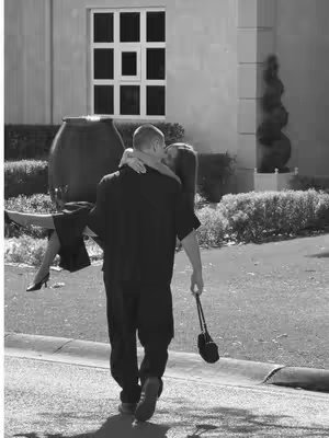 A black and white photo shows a man carrying a woman in his arms. The man is viewed from the back, walking towards the right. He is wearing dark clothing and holding a black purse. The woman is wearing a black outfit and high heels. The setting appears to be outside, with a building in the background and bushes along a walkway.