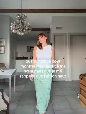 A woman stands in front of a kitchen area dressed in a white top and a long, shimmering turquoise skirt. The background includes a chandelier and other home decor.