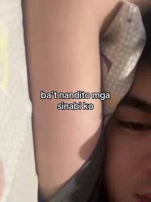 A selfie-style shot from a low angle shows a person lying in bed, with a portion of their arm and face visible. The person's arm is bent at the elbow and raised, with the forearm and hand obscured off-screen. The visible skin tone is fair. The person's shoulder and upper chest are partially visible. The background includes a patterned bedsheet and a patterned pillow.