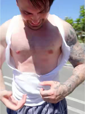 A man with dark hair is pulling down his white tank top, revealing his chest. He appears outdoors on a bright day.
