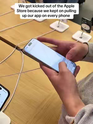 A shot from a first-person perspective, the scene reveals a person's hands holding a smartphone. The phone displays a white app screen with a touchscreen keyboard at the bottom. The hands are set against a light wood-toned surface. Wires and display stands are partially visible, suggestive of a retail or tech environment like an Apple Store. In the background, another phone sits on a display stand, and above the phone is the text overlay.